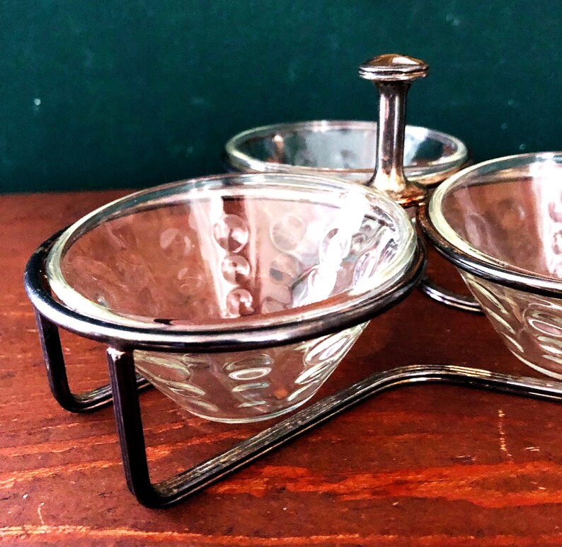 K&ouml;nnte beinhalten: Ein Vintage-Gew&uuml;rzset mit drei durchsichtigen Glasschalen mit strukturiertem Muster, die jeweils in einem silberfarbenen Metallrahmen gehalten werden. Ein zentraler Griff verbindet sich mit einer kleineren, runden Glasschale. Das Set steht auf einer Holzoberfl&auml;che.