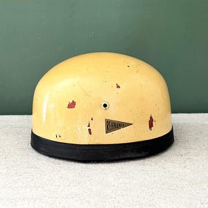 Peut inclure: Casque vintage crème avec une base noire. Le casque présente un petit trou et un emblème triangulaire avec l'inscription "REHNANIA". Des éclats de peinture rouge ajoutent à l'aspect vieilli. Cet objet de collection date probablement du milieu du XXe siècle.