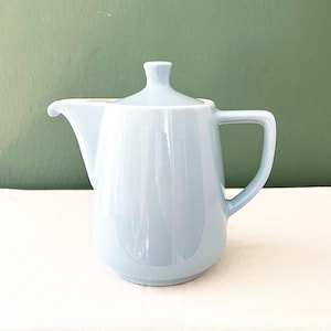 Puede incluir: Una tetera de cerámica de color azul claro con un acabado liso y brillante. La tetera presenta un cuerpo redondeado, un pico curvo, un asa resistente en el lado derecho y una tapa a juego con un pequeño pomo en la parte superior.
