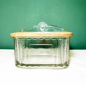 Op de afbeelding: Een heldere rechthoekige glazen container met een houten deksel. De container heeft een textuurpatroon aan de zijkanten en een doorzichtige handgreep aan de bovenkant. Het houten deksel is lichtbruin. De achtergrond is effen groen.