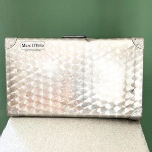 Pode incluir: Mala prateada com superfície texturizada com padrão de losangos. A mala tem cantos reforçados e uma pega. A marca "Marc O'Polo" é visível. Está sobre uma superfície branca contra um fundo verde.