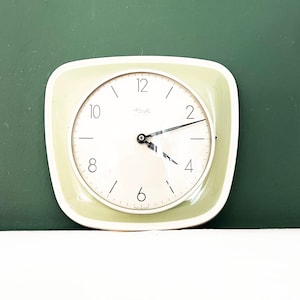 May include: A square wall clock with rounded corners featuring a light green frame and a white circular face. The clock has black numerals at the 2, 4, 6, 8, 10, and 12 positions, with thin black hour and minute hands and a thin black second hand.