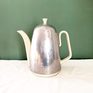 Op de afbeelding: Een vintage zilverkleurige theepot met een gehamerde textuur. De tuit, het handvat, het deksel en de basis zijn crèmekleurig, wat een tweekleurig effect creëert. De theepot staat tegen een groene achtergrond.