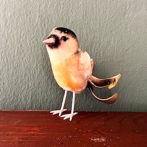 May include: A plush bird toy with a black head, yellow body, and pink legs. The bird has a small, pink beak and a yellow tag on its tail.