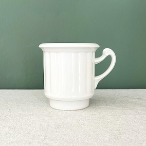 Könnte beinhalten: Eine weiße Keramiktasse mit Henkel, die vertikale Rippendetails am Körper aufweist. Die Tasse hat ein klassisches Design und steht auf einem leicht erhöhten Sockel. Der Hintergrund ist einfarbig grün.