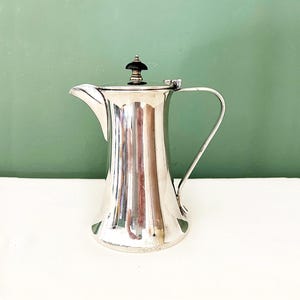 Puede incluir: Jarra plateada con un pico y asa curvados. La jarra tiene una base acampanada y una tapa con un pomo decorativo oscuro. La superficie refleja la luz, creando una apariencia brillante. El fondo es de color verde liso.