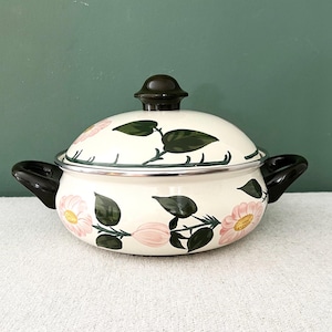Peut inclure: Une casserole en émail crème avec un couvercle assorti. Elle est ornée de fleurs roses et de feuilles vertes. Elle possède des poignées noires et un bouton noir sur le couvercle. Posée sur une surface blanche.