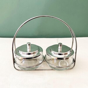 Op de afbeelding: Een heldere glazen kruidenset met twee ronde potten, elk met een zilveren deksel en een kleine lepel. De potten staan in een zilveren metalen houder met een handvat. De achtergrond is effen groen.
