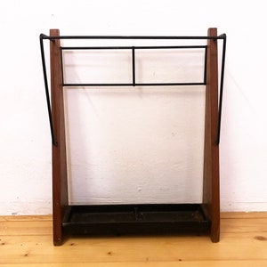 Puede incluir: Un paragüero vintage de madera oscura con detalles de metal negro. El soporte tiene una bandeja rectangular en la base y un estante para colgar paraguas. El diseño es típico de los muebles de mediados de siglo.