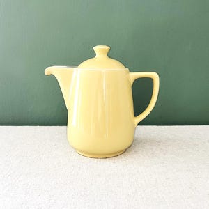 Może przedstawiać: Jasnożółty ceramiczny imbryk z pasującą pokrywką i zakrzywionym uchwytem. Imbryk ma dzióbek do nalewania i stoi na białej powierzchni na zielonym tle. Imbryk ma klasyczny design.