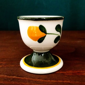 Puede incluir: Un huevo de cerámica blanca con un diseño floral verde y amarillo. La taza tiene una base ancha y una parte superior estrecha.