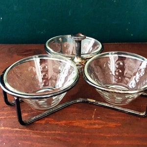 K&ouml;nnte beinhalten: Ein dreiteiliges Serviergeschirr mit durchsichtigen Glasschalen und einem silberfarbenen Metallrahmen. Die Schalen haben ein dekoratives Muster und werden von einem schwarzen Metallst&auml;nder gehalten. Die mittlere Schale hat einen Griff.