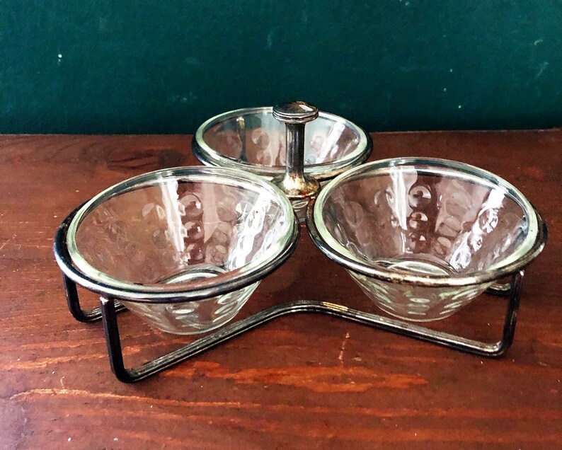 K&ouml;nnte beinhalten: Ein dreiteiliges Serviergeschirr mit durchsichtigen Glasschalen und einem silberfarbenen Metallrahmen. Die Schalen haben ein dekoratives Muster und werden von einem schwarzen Metallst&auml;nder gehalten. Die mittlere Schale hat einen Griff.