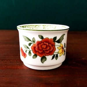 以下が含まれることがあります： オレンジ、黄色、緑の花が描かれた白い陶器の卵カップ。カップの縁には緑と白の幾何学模様が施されています。