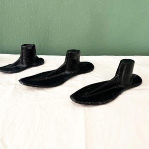 Puede incluir: Tres hormas de zapatos de metal negro, utilizadas para dar forma a los zapatos, sobre una superficie blanca.