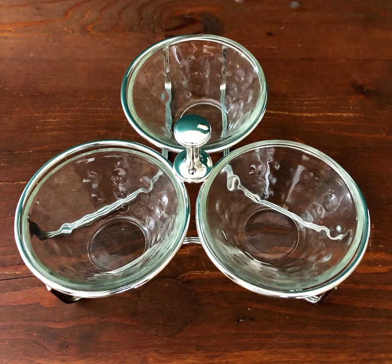 K&ouml;nnte beinhalten: Ein dreiteiliges Serviergeschirr mit durchsichtigen Glasschalen und einem silberfarbenen Metallrahmen. Die Schalen sind in einem dreieckigen Muster angeordnet, jede mit strukturiertem Innenraum. Der Rahmen hat ein dekoratives Design und einen zentralen Griff.