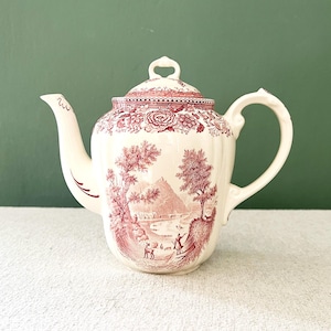 Puede incluir: Tetera de cerámica color crema con un diseño rojo. La tetera presenta una escena de paisaje con figuras, árboles y una montaña. Tiene un pico curvo, un asa de bucle y una tapa con un pequeño pomo. El diseño se repite en la tapa.