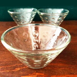 K&ouml;nnte beinhalten: Drei klare Glasschalen mit einem strukturierten, gepunkteten Muster. Die Schalen haben eine flache, konische Form, ideal zum Servieren kleiner Portionen. Die Schalen sind auf einer Holzoberfl&auml;che angeordnet.