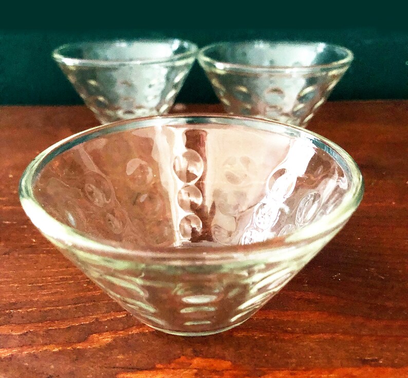 K&ouml;nnte beinhalten: Drei klare Glasschalen mit einem strukturierten, gepunkteten Muster. Die Schalen haben eine flache, konische Form, ideal zum Servieren kleiner Portionen. Die Schalen sind auf einer Holzoberfl&auml;che angeordnet.