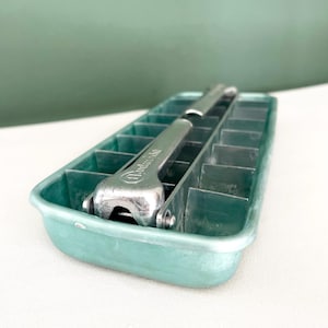 Pode incluir: Uma bandeja de cubos de gelo vintage turquesa com uma alavanca de liberação de cubos de gelo de metal prateado. A bandeja é retangular com compartimentos individuais para cubos de gelo. A alavanca é marcada com o texto "Handy-aide".