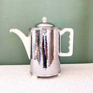 Könnte beinhalten: Eine silberfarbene Vintage-Kaffeekanne mit gehämmertem Muster. Sie hat einen weißen Griff, Ausguss und Deckelknopf. Die Kanne steht auf kleinen weißen Füßen und hat einen Deckel. Die Kanne ist etwa 20 cm hoch.