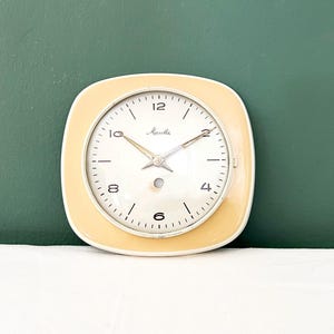 Può includere: Un orologio vintage color crema con quadrante quadrato e angoli arrotondati. Il quadrante è bianco con numeri e lancette nere. L'orologio ha la scritta "Mauthe" stampata sul quadrante.