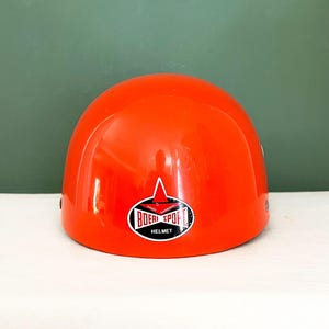 Può includere: Un casco protettivo arancione brillante con una finitura liscia e lucida. Al centro della parte anteriore si trova un logo ovale bianco e nero con una stella rossa e la scritta BOERI SPORT HELMET. Il casco ha una forma a cupola arrotondata.