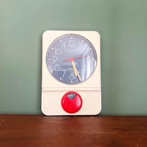 Op de afbeelding: Een crèmekleurige vintage klok met een donkerblauwe wijzerplaat en witte cijfers. De klok heeft rode en gele wijzers en een rode wijzerplaat aan de onderkant. De klok is aan een groene muur bevestigd.