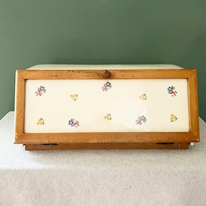 Könnte beinhalten: Holz-Brotdose mit Klappdeckel. Die Frontplatte hat einen cremefarbenen Hintergrund, der mit kleinen Blumenmustern in Blau, Gelb und Rosa verziert ist. Sie hat einen hellbraunen Holzrahmen und einen Holzknopf.