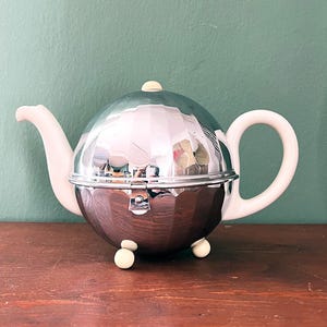 Könnte beinhalten: Eine silberne und weiße Teekanne mit rundem Korpus und Deckel. Die Teekanne hat einen weißen Henkel und Ausguss und steht auf drei weißen Füßen.