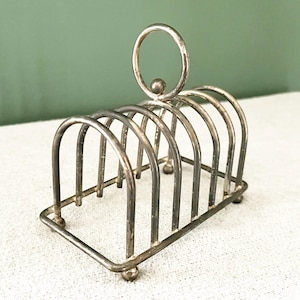 Puede incluir: Un tostador de metal plateado con seis ranuras arqueadas. Tiene una base rectangular y un asa con un lazo circular. El soporte está diseñado para sostener rebanadas de tostadas en posición vertical.