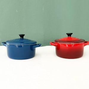 Op de afbeelding: Twee ronde geëmailleerde gietijzeren mini-cocottes met deksels. De ene is diepblauw en de andere is felrood. Beide hebben twee handgrepen aan de zijkant, een zwarte knop op het deksel en het in reliëf gemaakte Le Creuset-logo op het deksel.