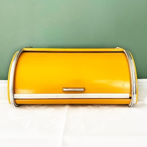 Può includere: Un portapane giallo brillante con coperchio a serrandina curvo. Il contenitore presenta una maniglia orizzontale in metallo argentato sulla parte anteriore e finiture in metallo argentato lucido lungo i bordi laterali, per un design funzionale.