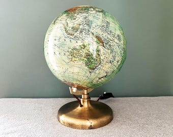 Op de afbeelding: Een vintage globelamp met een gedetailleerde wereldkaart. De globe is gemonteerd op een messingkleurige voet en standaard. De globe toont tinten groen, bruin en blauw. Een zwart netsnoer komt uit de basis.