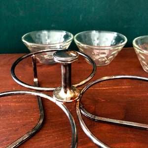 K&ouml;nnte beinhalten: Ein silberfarbener Metall-Serviertablett mit einem zentralen Griff und kreisf&ouml;rmigen Haltern f&uuml;r kleine Glasschalen. Die Schalen haben ein strukturiertes Muster. Das Tablett steht auf einer Holzoberfl&auml;che.