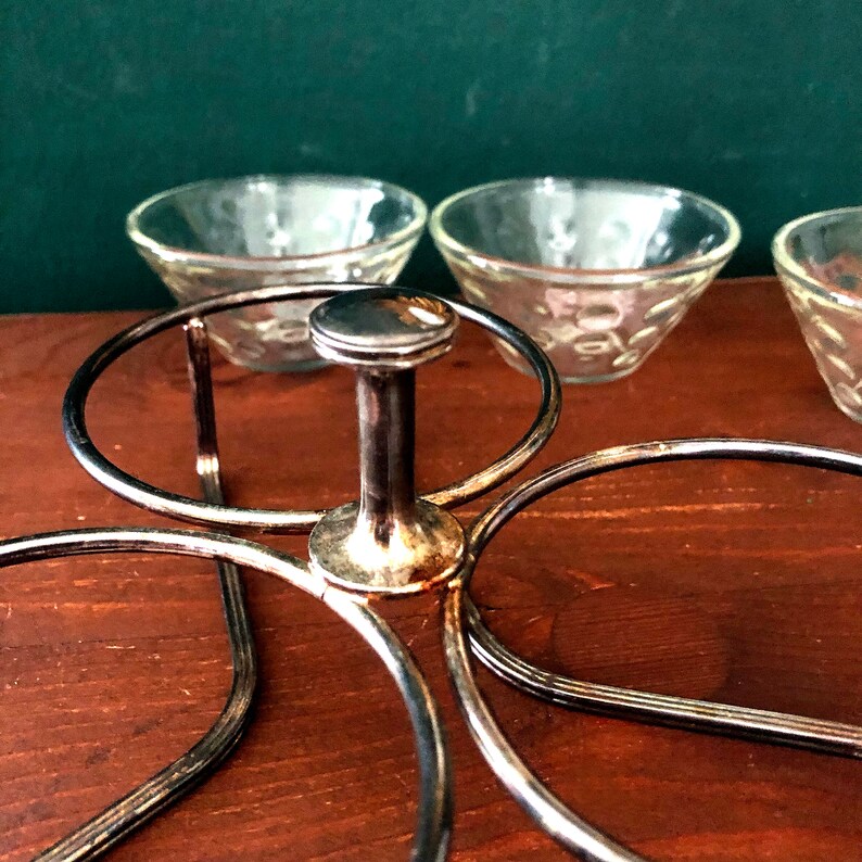 K&ouml;nnte beinhalten: Ein silberfarbener Metall-Serviertablett mit einem zentralen Griff und kreisf&ouml;rmigen Haltern f&uuml;r kleine Glasschalen. Die Schalen haben ein strukturiertes Muster. Das Tablett steht auf einer Holzoberfl&auml;che.