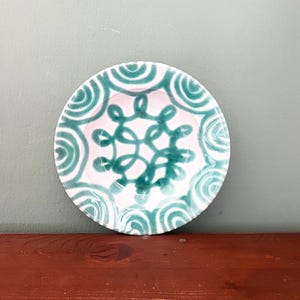 May include: A small, round ceramic plate with a white base and teal green decorative elements. The plate features a central teal green swirl design and a border of teal green semi-circles. The plate is on a wooden surface.