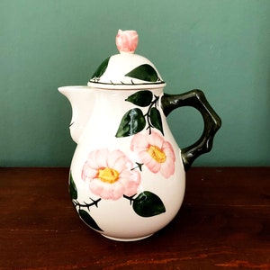 Könnte beinhalten: Eine weiße Teekanne aus Keramik mit einem Blumendesign. Die Teekanne hat einen Deckel mit einem Knopf in Form einer Blume. Die Teekanne hat einen Henkel und ist mit rosa Rosen und grünen Blättern verziert.