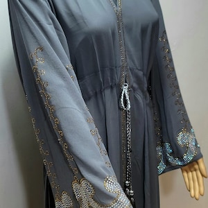 May include: A gray abaya with a hood and long sleeves. The abaya is decorated with a beaded design in a floral pattern. The abaya has a drawstring waist.