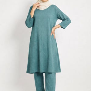 May include: A teal-colored two-piece outfit, consisting of a long-sleeved tunic and matching pants. The tunic has a flared silhouette and the fabric has a vertical ribbed texture. The outfit is worn with a cream-colored head covering.