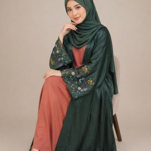 May include: A woman wearing a dark green satin abaya with floral embroidery on the sleeves, paired with a peach-colored dress and a matching green hijab. She is seated on a chair, with a neutral background.