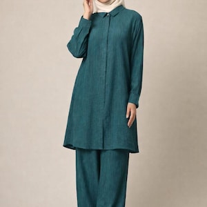 May include: Teal-colored, long-sleeved tunic and wide-leg pants set. The tunic has a collared neckline and button-down front. The outfit is made of a soft, flowing fabric. The model is wearing a white hijab.