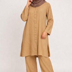 May include: A tan two-piece outfit featuring a long-sleeved tunic top and wide-leg pants. The tunic has a button-down front and side slits. The outfit is made of a textured fabric and is paired with a matching headscarf and sandals.