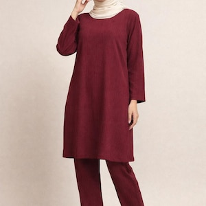 May include: A burgundy two-piece outfit, consisting of a long-sleeved tunic and matching pants. The tunic has a slightly A-line shape. The outfit is made of a textured fabric. The person in the image is wearing a cream-colored hijab.