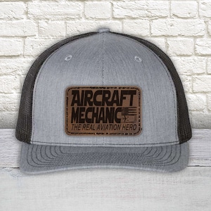 May include: Gray trucker hat with a black mesh back and a curved brim. A brown leather patch on the front displays the words "AIRCRAFT MECHANIC" and "THE REAL AVIATION HERO" with an American flag graphic.