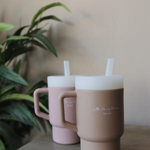 May include: Two silicone mugs with lids and straws. The mug on the left is pink and says "Daisy Co. Est 2020". The mug on the right is brown and says "Little Daisy Dream Est 2020".