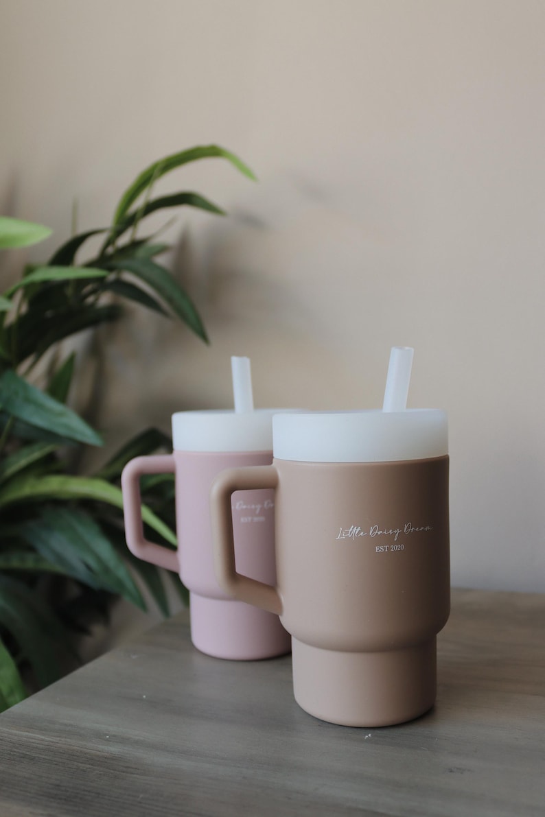 May include: Two silicone mugs with lids and straws. The mug on the left is pink and says "Daisy Co. Est 2020". The mug on the right is brown and says "Little Daisy Dream Est 2020".