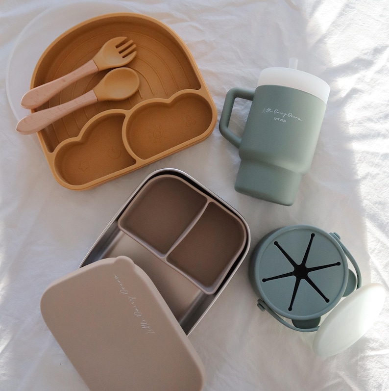 May include: A set of silicone baby feeding products in green and brown. The set includes a divided plate, a cup with a lid, a small container with a lid, and a set of utensils.