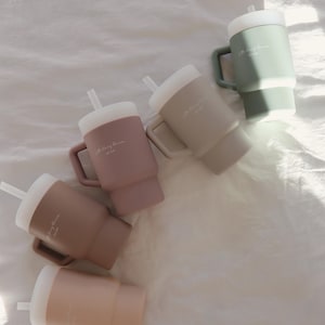May include: Five silicone travel mugs with lids and straws in various shades of brown, beige, and green. Each mug has the text "The Cozy Room" printed on the side.