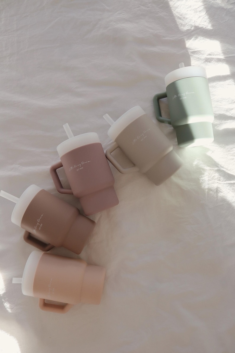 May include: Five silicone travel mugs with lids and straws in various shades of brown, beige, and green. Each mug has the text "The Cozy Room" printed on the side.